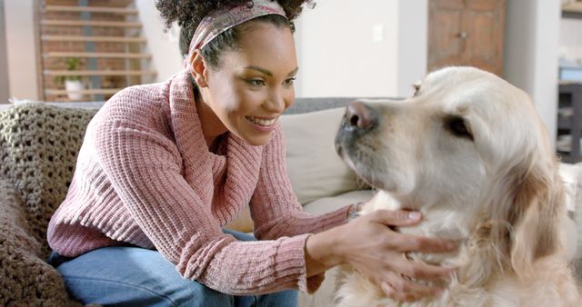 This image captures a cheerful woman with curly hair wearing a pink sweater, lovingly petting her Golden Retriever on a cozy couch at home. Perfect for use in articles and advertisements about pet care, home life, friendship, and human-animal bonding.