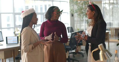 Diverse women coworkers chatting during office holiday party wearing festive accessories