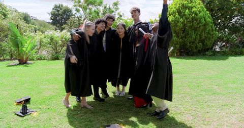 Diverse graduating students celebrating outdoors wearing black gowns holding diplomas