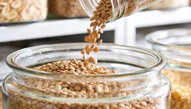 Pouring tan lentils from tilted jar into wide-mouthed glass storage jar on pantry shelf