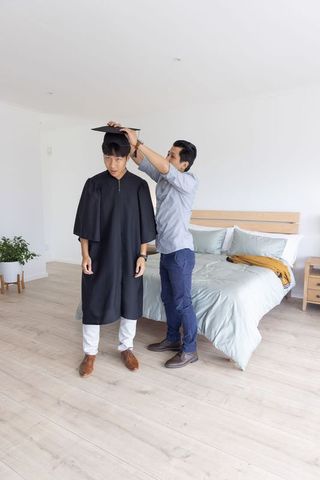 Asian graduate adjusting mortarboard with friend in bright minimalist bedroom