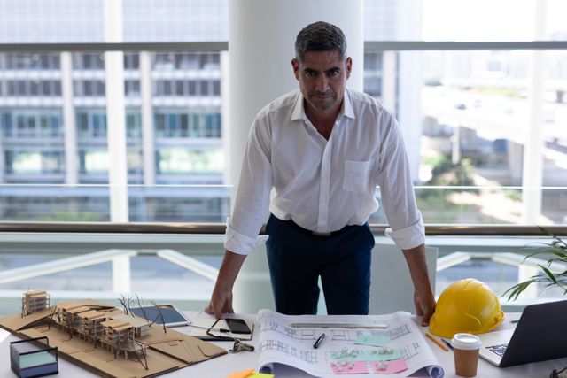 Mature male architect standing at desk in modern office, looking serious. He is surrounded by architectural models, blueprints, and a yellow hard hat, indicating a focus on construction and design. This image can be used for business, architecture, engineering, and professional work themes, highlighting dedication and expertise in the field.
