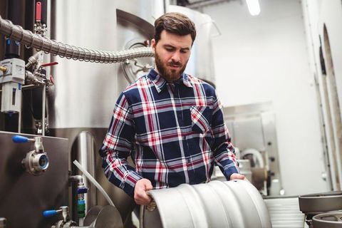 Brewery worker lifting metal keg amidst stainless steel tanks