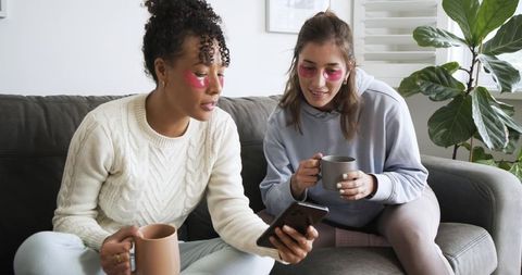 Two friends wearing under-eye patches sharing coffee and scrolling smartphone on cozy sofa