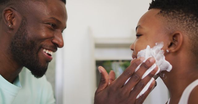 Father smiling while showing son how to shave using shaving cream in home bathroom. Perfect for illustrating family bonding moments, parenting tips, grooming tutorials, and everyday family life.