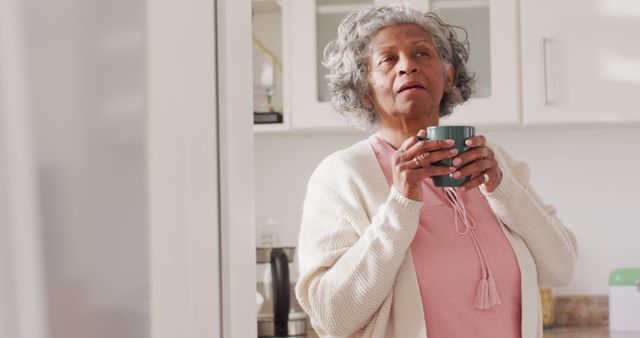 Senior woman in casual clothing drinking coffee in her kitchen, looking contemplative. Ideal for use in lifestyle blogs, health articles, and advertisements highlighting senior wellness and peaceful mornings at home.