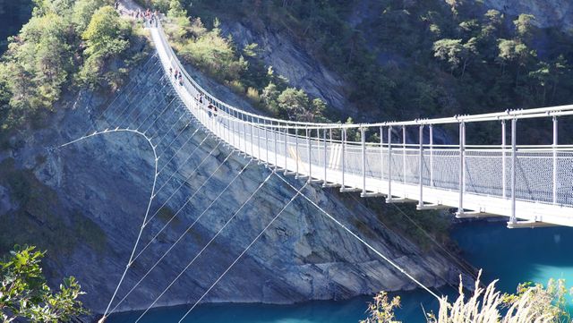 Ideal for usage in travel guides, outdoor adventure promotions, hiking advertisements, and nature documentaries. Depicts a suspension bridge stretched over a picturesque mountain gorge with clear blue water, surrounded by lush greenery and rugged rocks, emphasizing the beauty and thrill of outdoor exploration.