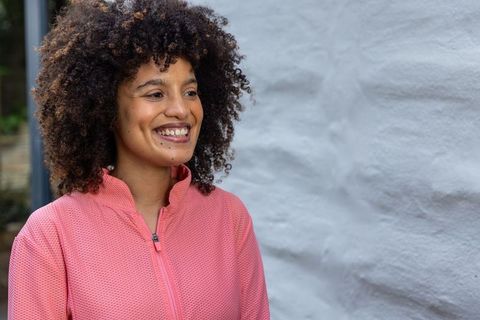 Smiling african american woman wearing pink zip-up jacket in courtyard with stucco wall