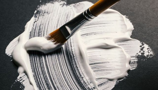 Flat brush spreading thick white paint on dark board creating glossy textured ridges