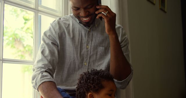 Father happily talking on phone while sitting by window, holding young child on lap. Perfect for themes of family, parenting, work-life balance, communication, and home life. Use in articles on fatherhood, happy family moments, or work-from-home dynamics.
