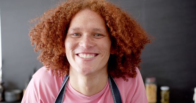 Young person with curly red hair smiling warmly and wearing pink shirt with apron. Ideal for representations of happiness, friendliness, and positive emotions in websites, marketing materials, and social media. Fits themes like diversity, self-expression, and everyday joy.
