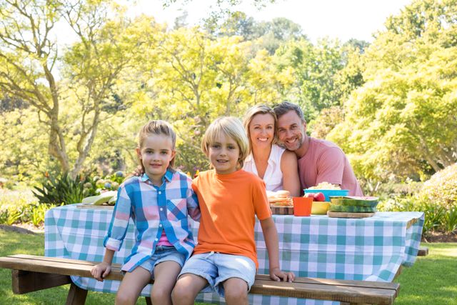 Family enjoying a sunny day at a park picnic, sitting at a table with various foods. Ideal for content related to family bonding, outdoor activities, healthy lifestyles, and recreational activities. Can be used for promotional materials for parks, family events, and advertisements for picnic products.