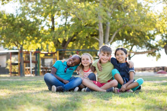 Children enjoying a sunny day in the park, sitting together on the grass with arms around each other. Perfect for themes related to friendship, childhood, outdoor activities, and diversity. Ideal for use in educational materials, advertisements for children's products, and community event promotions.