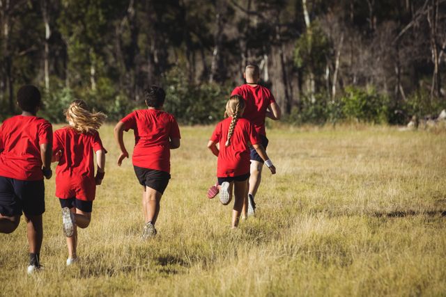 A trainer is leading a group of kids in a boot camp training session outdoors on a sunny day. The children are running in a grassy field surrounded by trees. This image can be used for promoting fitness classes, outdoor activities for children, health and wellness programs, team building exercises, and educational materials on physical education.
