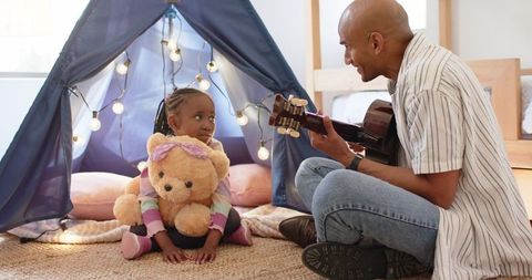 Father playing guitar with african american daughter holding teddy bear in lit teepee bedroom
