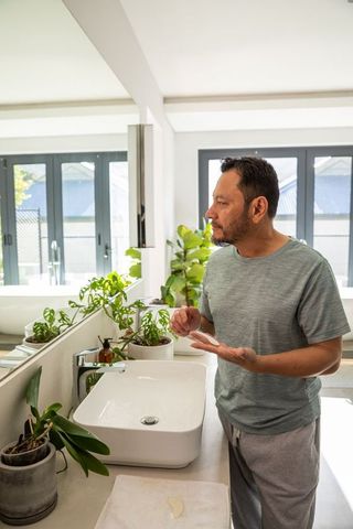 Asian man applying skincare at vanity in modern minimalist bright bathroom with plants