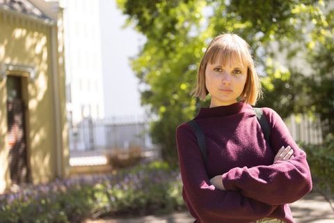 Young woman standing with arms folded in sunlit courtyard wearing maroon sweater
