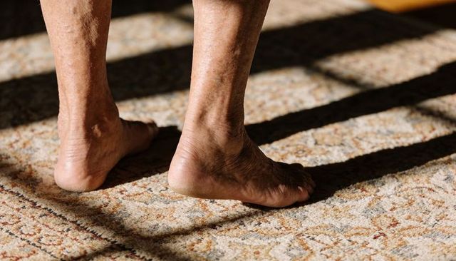 Sunlit senior feet lifting heel on patterned carpet with blinds casting geometric shadows