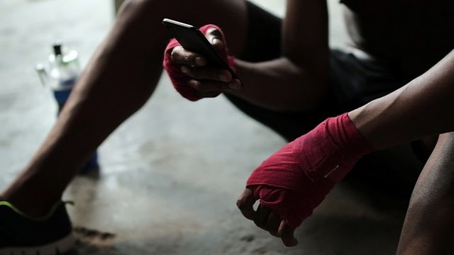 Dark room scene captures a fitness enthusiast relaxing post-exercise while interacting with his phone. The image showcases red hand wraps, sporty attire, and a water bottle. Useful for themes related to modern fitness routines, digital interactivity in sports, or athletic lifestyle promotions. Ideal for blogs, fitness apps, or advertisements targeting health and tech-savvy audiences.