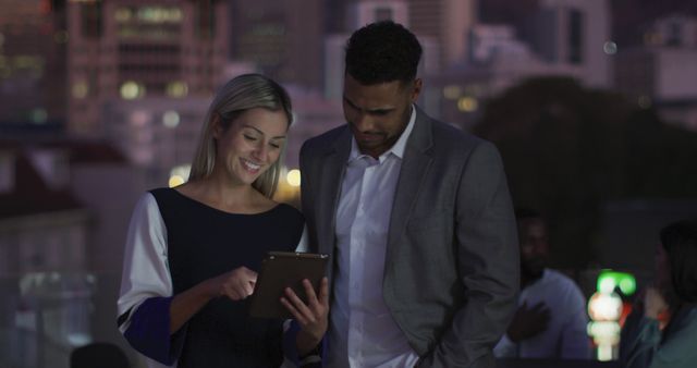 This stock photo shows two colleagues, a woman and a man, reviewing business plans on a tablet during an evening rooftop meeting. The woman is smiling while pointing at the tablet screen, indicating engagement and productivity. The man is attentively looking at the screen, dressed in a professional suit, suggesting a formal yet collaborative work environment. An urban background with buildings lit up hints at a city setting. Ideal for use in workplace culture, teamwork, urban work environment, professional collaboration, and technology in business imagery.