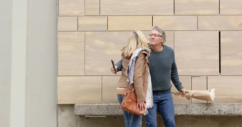 Mature couple sharing quick kiss beside modern stone wall holding bouquet and phone