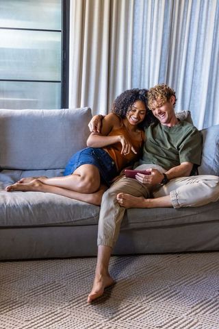 Cozy diverse couple relaxing on sofa laughing while sharing content on smartphone