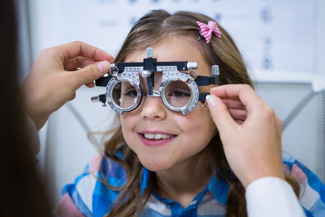 Young girl smiling while undergoing an eye examination with a trial frame in an ophthalmology clinic. Ideal for use in healthcare, pediatric optometry, vision care, and medical checkup contexts. Can be used in articles, brochures, and websites related to eye health and children's healthcare services.