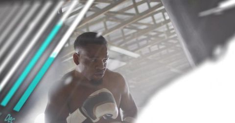 African american boxer training with heavy bag in industrial gym, backlit gritty workout