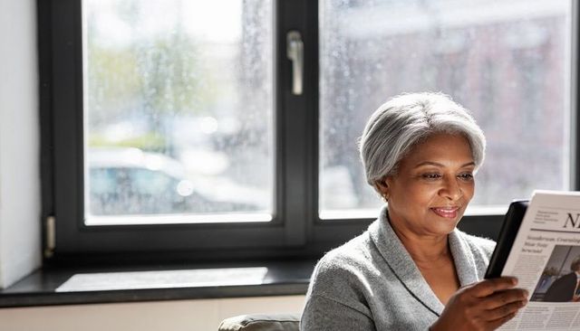 Senior woman reading newspaper by rainy window, cozy cardigan, natural morning light