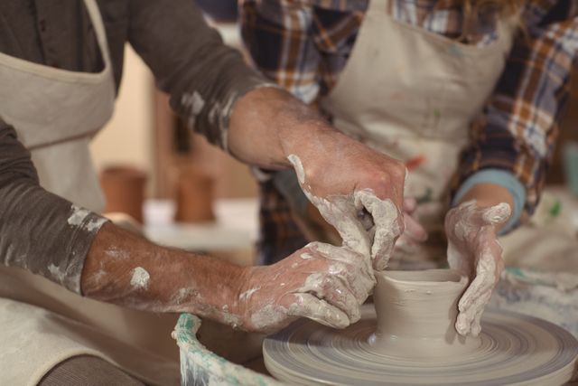 Image of a male potter assisting a female potter as they work together on a pottery wheel in a workshop. Both are wearing aprons, and their hands are covered in clay, demonstrating the hands-on nature of pottery making. This image is suitable for use in articles or advertisements related to art classes, craft workshops, pottery techniques, teamwork in creative activities, or hobbies.