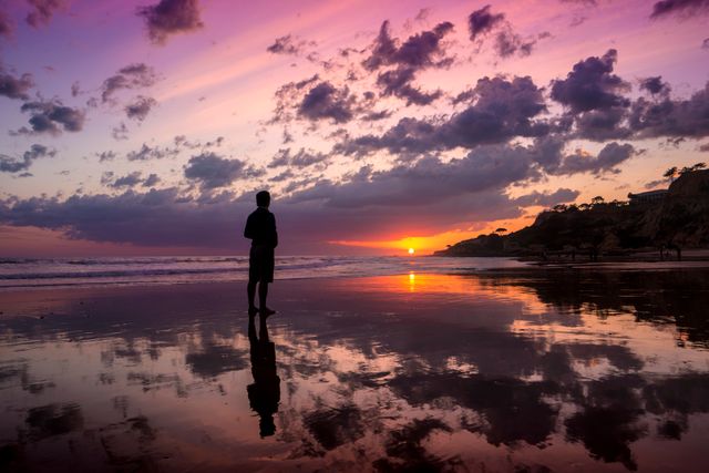 Person standing on wet beach watching vibrant sunset. Suitable for themes of reflection, tranquility, beach vacations, nature's beauty, peaceful getaways, and romantic evening settings.