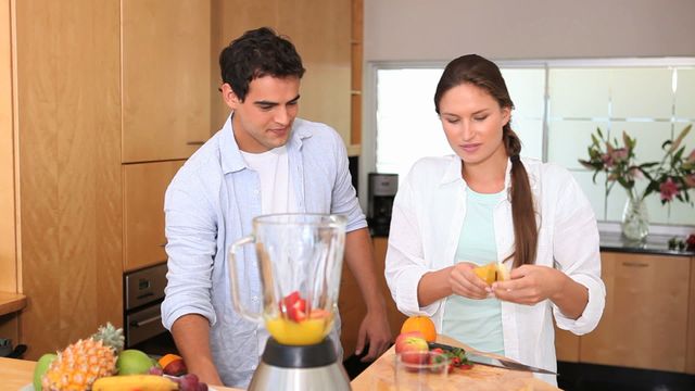 Young couple is preparing a fresh smoothie in a bright, modern kitchen. They are using a blender with various fresh fruits on the counter, showing a healthy lifestyle. Perfect for use in advertisements or articles about healthy living, wellness, nutritious recipes, cooking together, and family bonding.