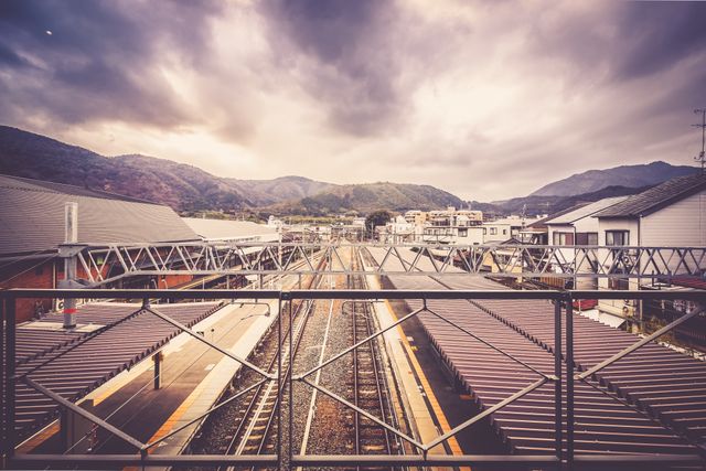 A calm train station with empty platforms against a backdrop of dramatic clouds and mountain scenery. The overhead crosswalk and urban surroundings add depth to the scene. Ideal for use in travel brochures, transportation advertisements, or generally to illustrate serene, rural settings in Japan.