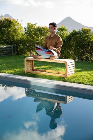Asian man meditating barefoot on wooden bench by pool in sunlit backyard mountain view