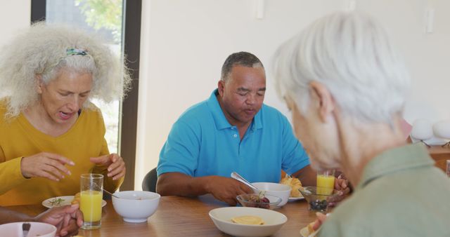 Senior friends share a cheerful morning together, having a breakfast of orange juice and fruit salad. The image is ideal for use in promotional materials for retirement homes, healthy lifestyle campaigns, and social gatherings for seniors. It highlights themes of companionship, enjoying life after retirement, and eating a nutritious meal.