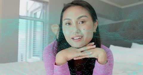 Contemplative asian woman leaning forward with cyan hologram overlay in pastel bedroom