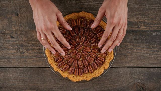 Hands arranging pecan halves on flaky pie crust flatlay over rustic wooden table