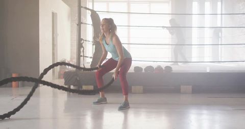 Woman swinging heavy battle ropes in boxing gym training with backlit ring and silhouettes