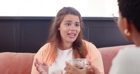 Young latina woman talking openly with black friend on cozy pink sofa during heartfelt conversation
