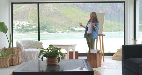 Mature artist painting in sunlit coastal living room studio with panoramic windows