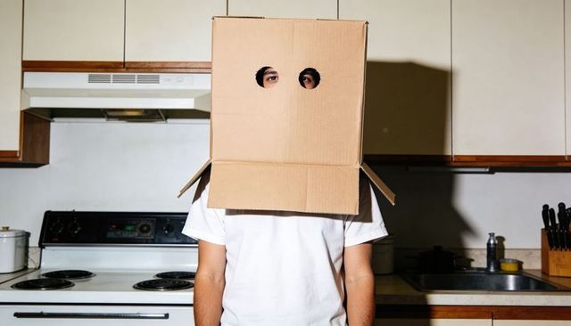 Anonymous person wearing cardboard box mask with cutout eyes standing in minimal kitchen