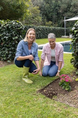 Middle-aged women planting pink flowers by pool, gardening together, backyard friendship