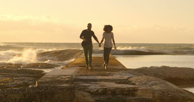 Romantic diverse couple holding hands and running on pier at sunrise, copy space. Summer, vacation, romance, love, relationship, free time and lifestyle, unaltered.