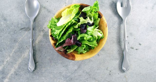 Freshly prepared green salad in a wooden bowl placed on a gray concrete background, with a fork and spoon beside it. This image is versatile for use in menus, websites, healthy eating campaigns, nutritional blogs, and dietary planning content. The clean and modern presentation highlights healthy and natural dietary choices.