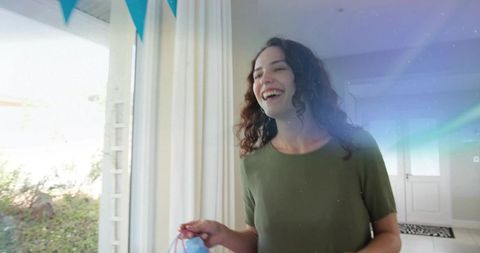 Smiling young woman holding pastel gift bag by sunlit door, bunting celebration at home