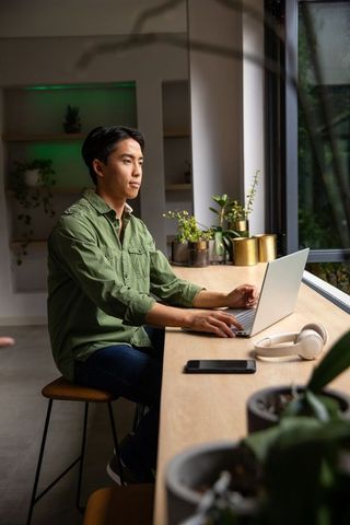 Focused asian man working on laptop at plant-lined window counter for remote productivity