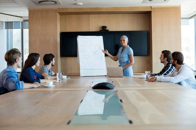 Woman presenting to her colleagues in a modern office conference room. She is standing next to a whiteboard with diagrams, engaging the team in discussion. Ideal for use in business, corporate training, teamwork, leadership, and professional development contexts.