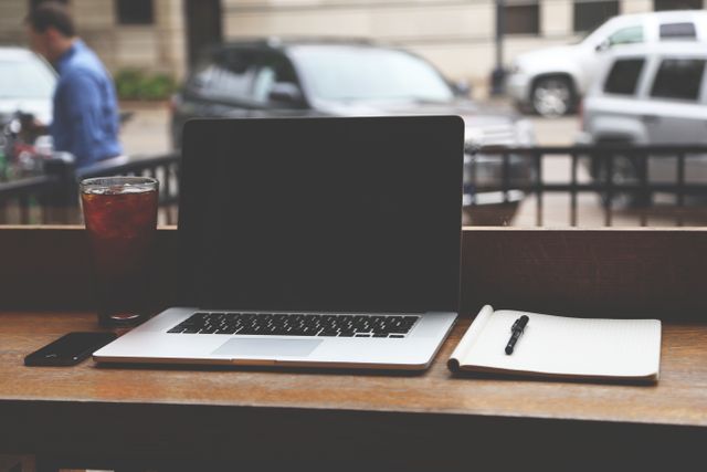 Open laptop next to a notebook and pen on a wooden table by a café window. A glass of iced tea is also visible. Cars and street blur seen outside. Ideal for themes on remote work, productivity, and casual work environment.