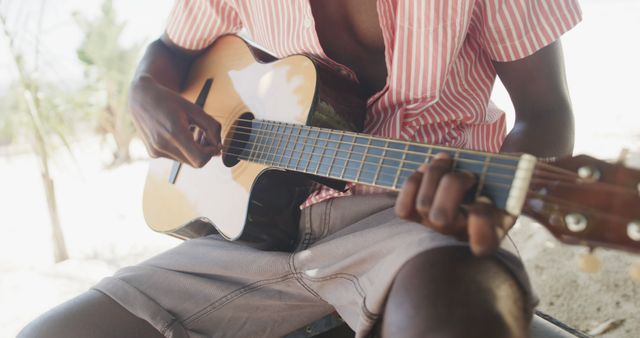 Midsection of african american man sitting playing guitar on sunny beach, slow motion. Summer, hobbies, relaxation and vacations.