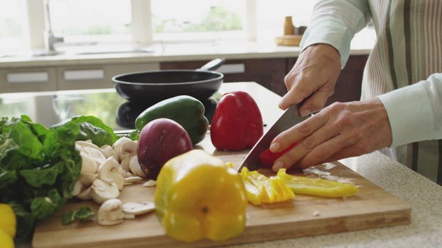 Senior woman slicing yellow and red bell peppers on wooden chopping board in modern kitchen. Bright, well-lit environment with clean surfaces and various fresh vegetables. Use to highlight healthy eating, home cooking, senior lifestyle, or food preparation during quarantine. Suitable for articles or promotions about healthy lifestyles, home activities, and senior living.